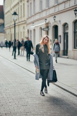 Beyaz, güzel, sarışın, uzun sarı saçlı, sokakta yürüyen, sırt çantası ve buketi tutan genç bir kadın. Gri paltolu, koyu yeşil kısa elbiseli ve siyah taytlı güzel, şık sarışın kadın şehir sokaklarında yürüyor..