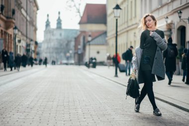 Beyaz, güzel, sarışın, uzun sarı saçlı, sokakta yürüyen, sırt çantası ve buketi tutan genç bir kadın. Gri paltolu, koyu yeşil kısa elbiseli ve siyah taytlı güzel, şık sarışın kadın Krakow şehrinde yürüyor.