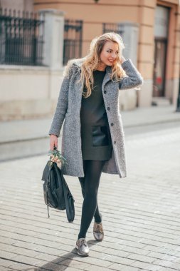 Sarışın, uzun ve dalgalı saçlı genç bir kadın sokaktaki insanlara gülümsüyor ve sarı saçlarını, sırt çantasını ve buketini tutuyor. Gri paltolu, koyu yeşil kısa elbiseli ve siyah taytlı güzel sarışın kadın şehir sokaklarında yürüyor..