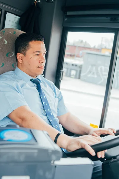 young hispanic bus driver is holding a wheel, looking at the road. latin man wears a blue shirt in bus. man is driving a bus. Transportation in the city.