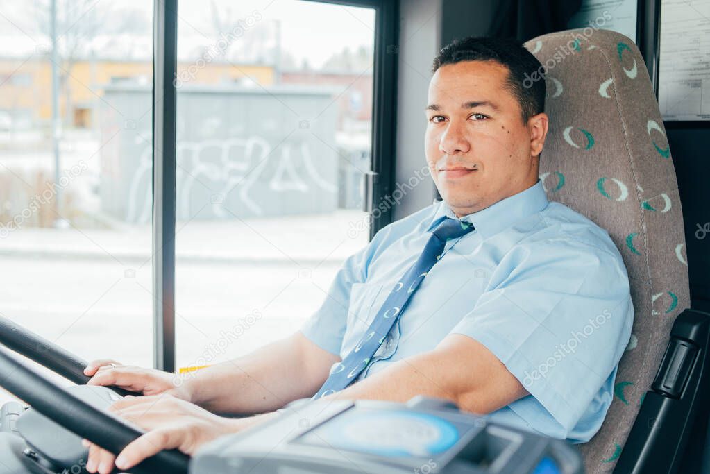 young hispanic bus driver is holding a wheel, looking at the road. latin man wears a blue shirt in bus. man is driving a bus. Transportation in the city.