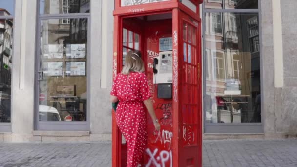 Młoda kobieta w czerwonej sukience zbliża się do czerwonej budki telefonicznej i używa miejskiego telefonu — Wideo stockowe