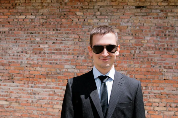 Fiatal férfi fekete öltöny, nyakkendő és a a téglafal háttér fekete napszemüveg — Stock Fotó