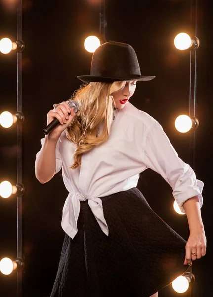 Jeune blonde jolie femme en chemise blanche, papillon, chapeau noir avec microphone chantant sur scène — Photo