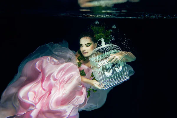 Víla nádherná mladá žena v růžových šatech s klecí v jejích rukou pod vodou — Stock fotografie