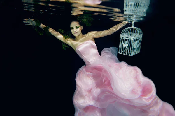 Fairy prachtige jonge vrouw in roze jurk met kooi in haar handen onderwater — Stockfoto