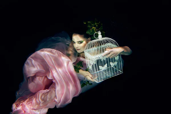 Víla nádherná mladá žena v růžových šatech s klecí v jejích rukou pod vodou — Stock fotografie