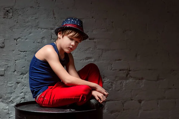 Портрет крутого молодого хлопчика хіп-хопу в блакитному капелюсі і червоних штанях, що сидять на чорному бочці в горищі — стокове фото