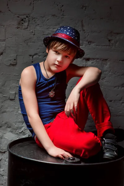 Портрет крутого молодого хлопчика хіп-хопу в блакитному капелюсі і червоних штанях, що сидять на чорному бочці в горищі — стокове фото