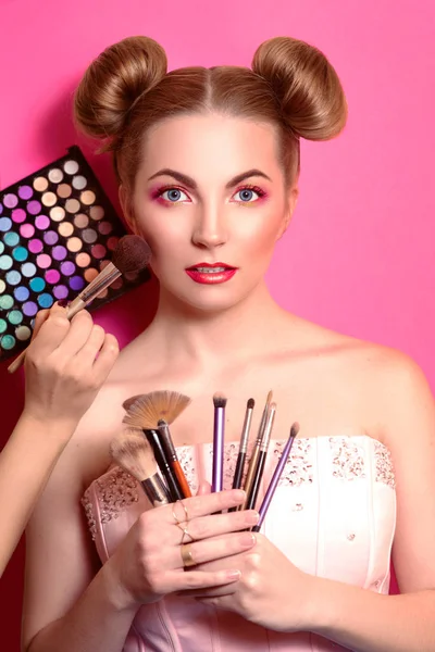 Younge attractive blonde woman with colorful make up with cosmetic brushes and shadows in her and make up master's hands