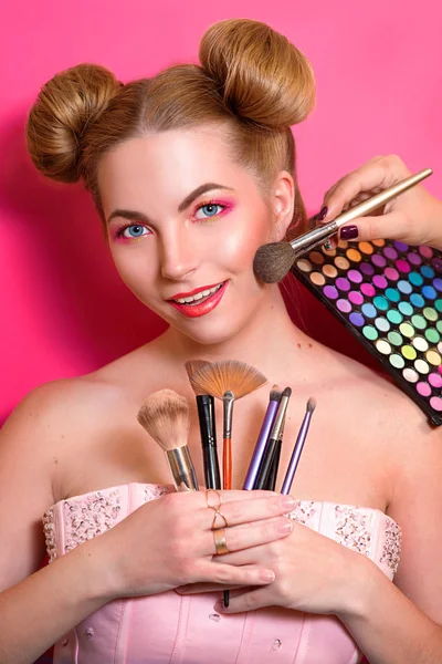 Younge attractive blonde woman with colorful make up with cosmetic brushes and shadows in her and make up master\'s hands