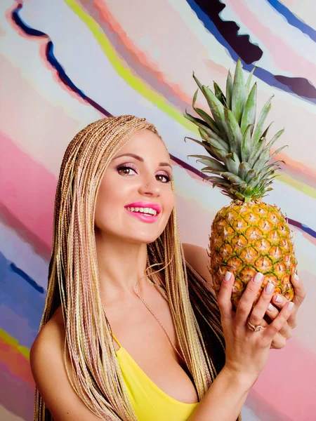 Jonge, leuke, mooie slanke blonde vrouw met Afrikaanse vlechten en met ananas in haar handen in gele jurk op kleurrijke achtergrond — Stockfoto
