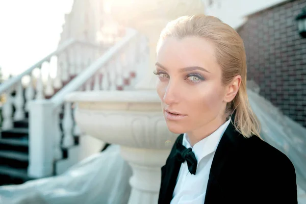 Retrato de elegante mulher loira bonita na moda no homem terno preto com laço ao ar livre quase fonte — Fotografia de Stock