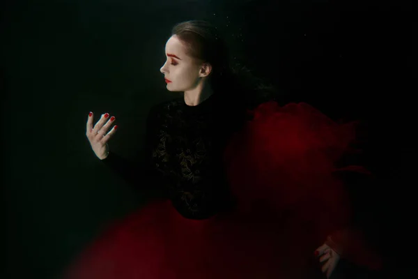 濃い色の背景には、スイミング プールに洋服の赤と黒のドレスの若い美しい女性の水中の肖像画 — ストック写真