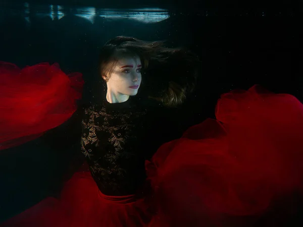 underwater portrait of young beautiful woman in black dress with red clothe in the swimming pool on the dark background