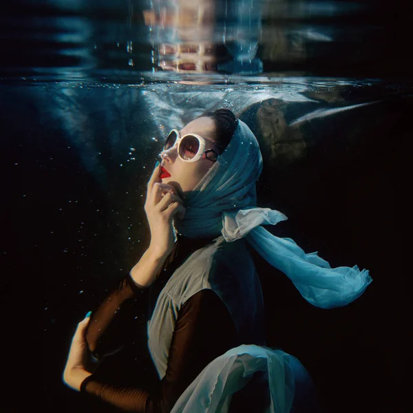 Retrato de atractiva joven morena en gafas de sol y bufanda azul bajo el agua en la piscina sobre fondo oscuro —  Fotos de Stock