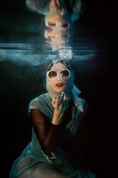 Potret wanita muda berambut cokelat yang menarik dengan kacamata hitam dan syal biru di bawah air di kolam renang dengan latar belakang gelap — Stok Foto