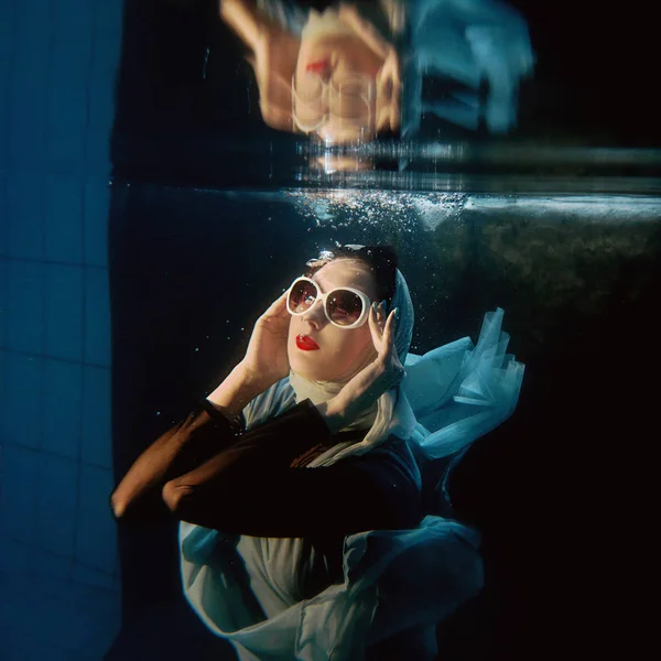 Portrait de jolie jeune femme brune en lunettes de soleil et écharpe bleue sous l'eau dans la piscine sur fond sombre — Photo