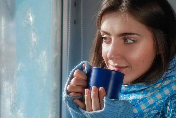Common smiling dreaming girl in sweater with the cup near the frozen window in the winter