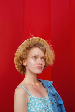 Hasır şapkalı, mavi elbisesi ve koyu kırmızı arka planda duran kot ceketli, kıvırcık saçlı genç bir kadın. Eğlence, yaz, moda, gençlik kavramı