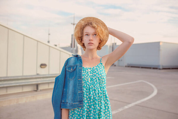 young curly redhead woman in straw hat, blue sundress and jeans jacket standing on the modern roof. Fun, summer, fashion, shooting, travel, youth concept. Copy spase