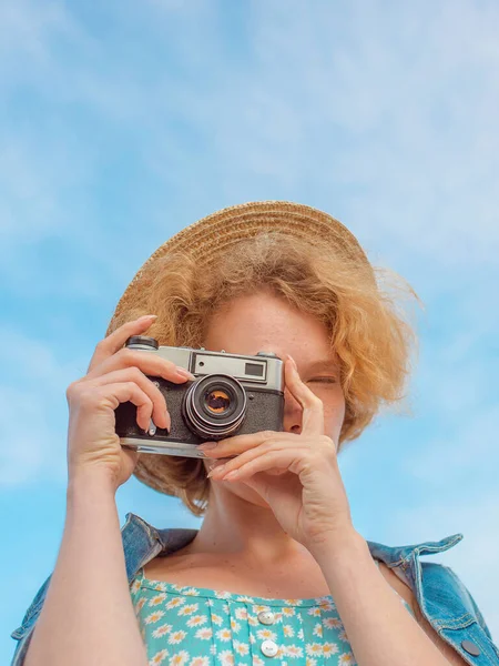 年轻的卷曲红头发的女人 戴着草帽 穿着蓝色的太阳衣和牛仔夹克 站在老式相机前 在蓝天的背景上拍照 青春的概念 — 图库照片