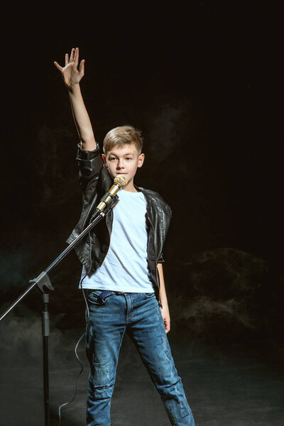 Портрет кавказского подростка в белой футболке, синих джинсах и кожаной куртке с микрофоном, поющим на темном фоне. Хобби и понятие славы
