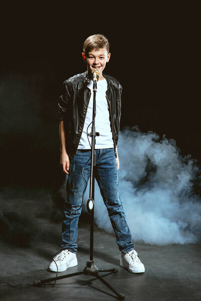 Портрет кавказского подростка в белой футболке, синих джинсах и кожаной куртке с микрофоном, поющим на темном фоне. Хобби и понятие славы
