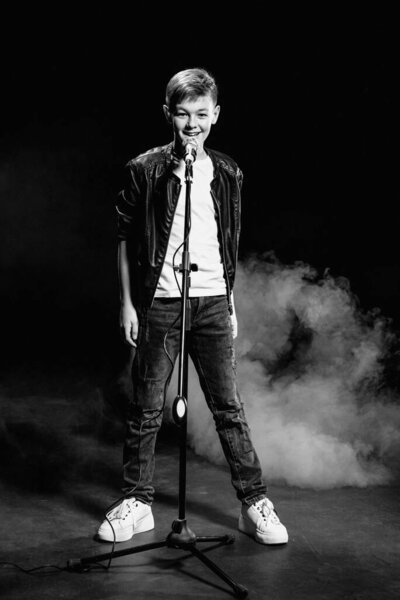 Монохромный портрет кавказского подростка в белой футболке, синих джинсах и кожаной куртке с микрофоном, поющим на темном фоне. Хобби, популярность и слава
