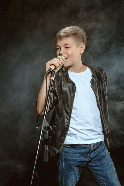 Портрет кавказского подростка в белой футболке, синих джинсах и кожаной куртке с микрофоном, поющим на темном фоне. Хобби и понятие славы
