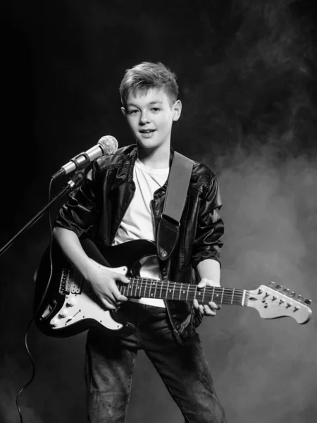 Monochrome portrait of caucasian teenager in white t-shirt, blue jeans and leather jacket with microphone singing on dark background. Hobby, popularity and glory concept