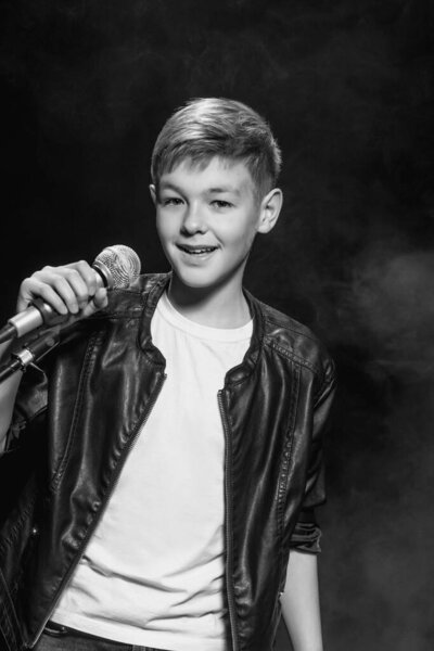 Монохромный портрет кавказского подростка в белой футболке, синих джинсах и кожаной куртке с микрофоном, поющим на темном фоне. Хобби, популярность и слава
