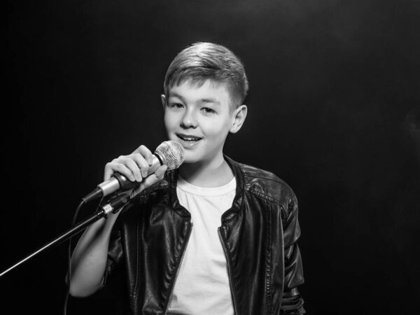 Монохромный портрет кавказского подростка в белой футболке, синих джинсах и кожаной куртке с микрофоном, поющим на темном фоне. Хобби, популярность и слава
