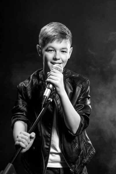 Монохромный портрет кавказского подростка в белой футболке, синих джинсах и кожаной куртке с микрофоном, поющим на темном фоне. Хобби, популярность и слава
