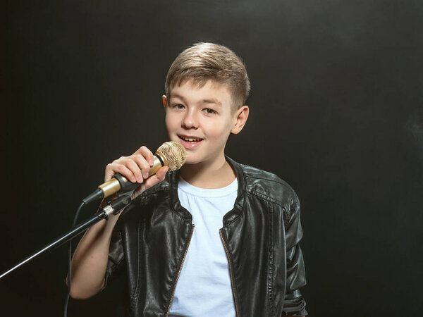 Портрет кавказского подростка в белой футболке, синих джинсах и кожаной куртке с микрофоном, поющим на темном фоне. Хобби и слава
