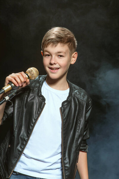 Портрет кавказского подростка в белой футболке, синих джинсах и кожаной куртке с микрофоном, поющим на темном фоне. Мбаппе, Караоке и концепция славы
