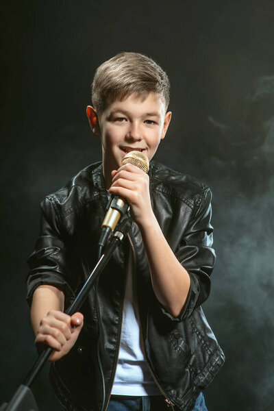 Портрет кавказского подростка в белой футболке, синих джинсах и кожаной куртке с микрофоном, поющим на темном фоне. Хобби и понятие славы
