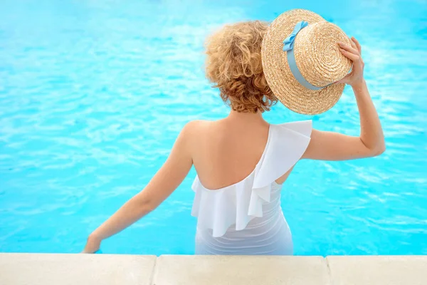 beautiful redhead (ginger) woman's back in the white swimming suit sitting by the swimming pool (or sea or ocean) in the straw hat in summer day. Summer, relax, wellness, recreation concept