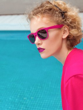 Fuşya ceketli, şık, kıvırcık saçlı, şık güneş gözlüklü ve yüzme havuzunun yanında siyah külotlu fevkalade bir kızın portresi. Moda, tarz, eğlence, eğlence, seyahat, makyaj konsepti. Film tahılı