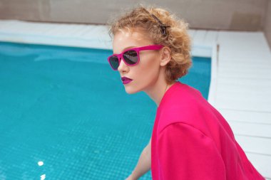 Fuşya ceketli, şık, kıvırcık saçlı, şık güneş gözlüklü ve yüzme havuzunun yanında siyah külotlu fevkalade bir kızın portresi. Moda, tarz, eğlence, eğlence, seyahat, makyaj konsepti. Film tahılı