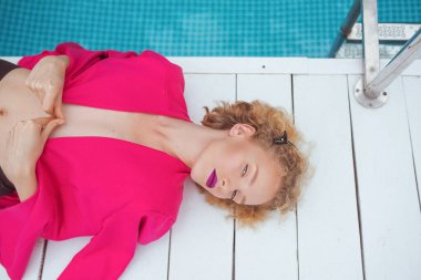 Havuzun yanında fuşya ceketli, şık, kıvırcık saçlı, genç bir kadının portresi. Moda, tarz, eğlence, eğlence, seyahat, makyaj konsepti. Film tahılı