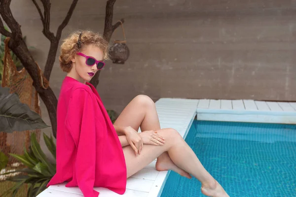 Fuşya ceketli, şık, kıvırcık saçlı, şık güneş gözlüklü ve yüzme havuzunun yanında siyah külotlu fevkalade bir kızın portresi. Moda, tarz, eğlence, eğlence, seyahat, makyaj konsepti. Film tahılı