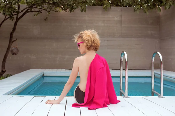 Fuşya ceketli, şık, kıvırcık saçlı, şık güneş gözlüklü ve yüzme havuzunun yanında siyah külotlu fevkalade bir kızın portresi. Moda, tarz, eğlence, eğlence, seyahat, makyaj konsepti. Film tahılı