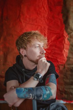 Kırmızı arka plandaki sandalyede oturan, elinde dövme olan kızıl saçlı bir adamın portresi. Sakin, moda, dövme, tarz kavramı. Film tahılı