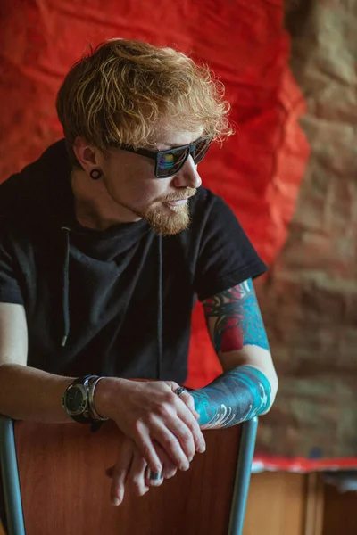 portrait of redhead stylish man with tattoo on his hand on the red background. Calm, fashion, tattoo, style concept. Film grain