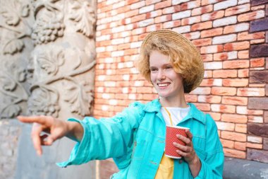 Mavi kot ceket ve hasır şapkalı (gezgin) neşeli, kıvırcık saçlı, genç bir kadın elinde kahve fincanı tutuyor ve işaretler karşısında şaşırıyor. Eğlence, yaz, seyahat, moda, gençlik kavramı