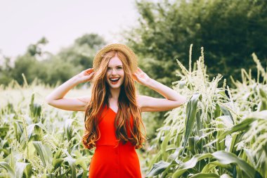   red-haired girl with freckles in red dress walking on a field of corn she is smiling a  sweet smile on her head Summer panama clipart