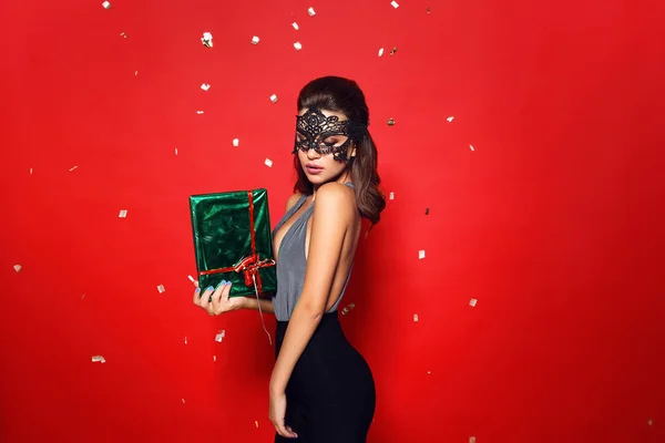 Beauty model woman wearing venetian black masquerade carnival mask at party over holiday red  background with magic stars.Christmas and New Year celebration.Glamour lady. holding a green gift