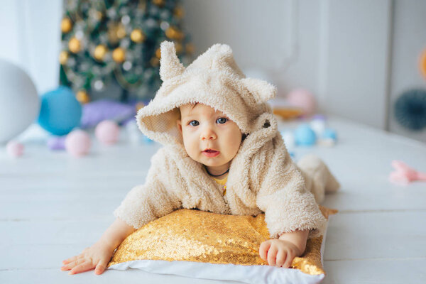 Beautiful little girl dressed in a funny suit lying on a pillow next to the gold-colored Christmas trees