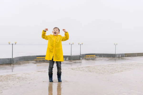 A happy boy in a yellow raincoat is standing on a wet city emban